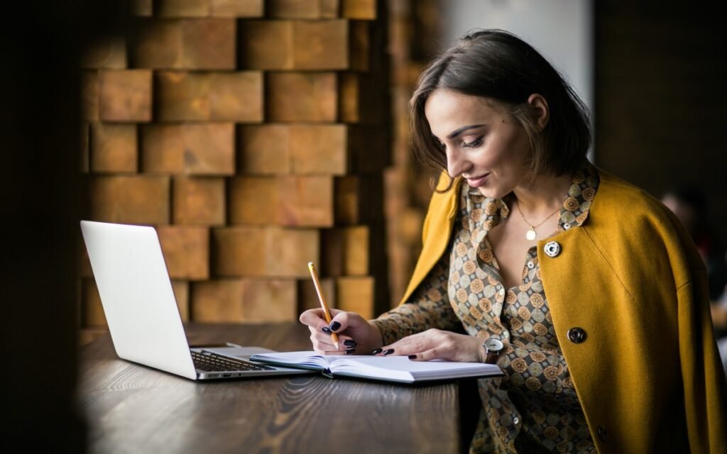 Woman working on a laptop and taking notes in a notebook.